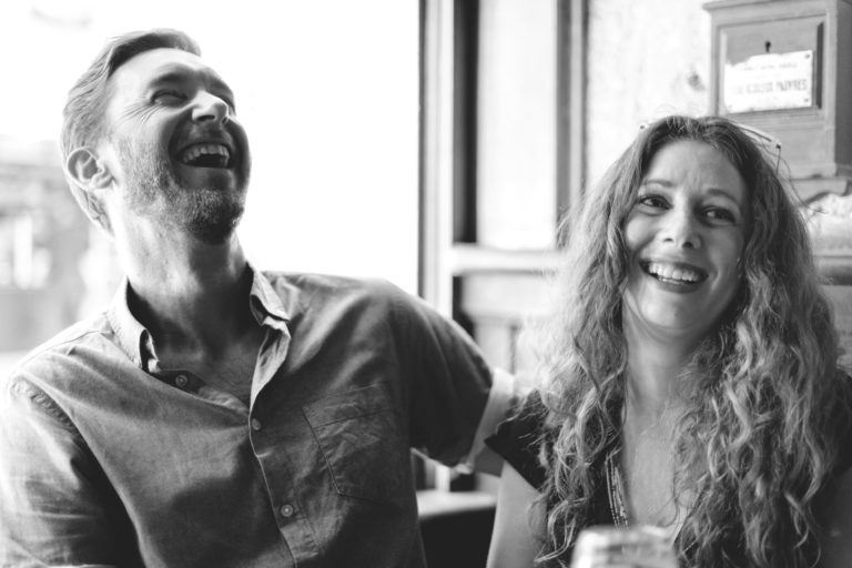 Une photo en noir et blanc capture un moment de joie et de coup de foudre au café Brun Bordeaux, avec un homme barbu en chemise boutonnée et une femme portant un collier, ses longs cheveux bouclés tombant en cascade alors qu'ils rient ensemble. Ils sont assis à l'intérieur près d'une fenêtre, une lumière douce illuminant leurs visages.