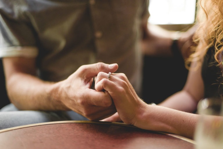 Deux personnages assis côte à côte, se tenant la main, de part et d'autre d'une table. Une main porte une bague au quatrième doigt, suggérant des fiançailles ou un mariage. Le décor semble intime, peut-être dans un café ou un restaurant comme le Café Brun Bordeaux. Seuls les torses et les bras des individus sont visibles, mettant l'accent sur le geste de se tenir la main.