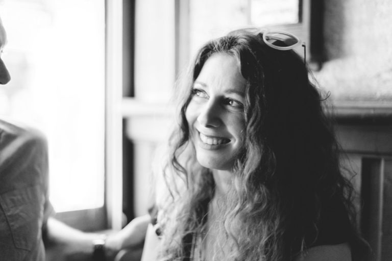 Une femme aux cheveux longs et bouclés, portant des lunettes de soleil sur la tête, sourit tout en regardant légèrement vers la gauche. La photo en noir et blanc la montre assise à l'intérieur près d'une fenêtre dans un endroit pittoresque rappelant Coup de foudre au café Brun Bordeaux, avec la lumière naturelle illuminant son visage. L'arrière-plan est légèrement flou, attirant l'attention sur son expression joyeuse.