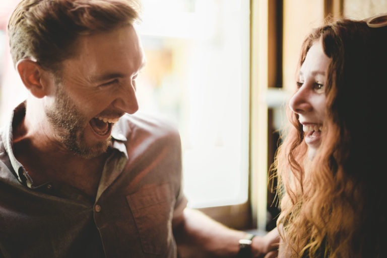 Un homme et une femme rient joyeusement ensemble, partageant un moment de joie qui rappelle un coup de foudre au café. L'homme, aux cheveux bruns courts et à la barbe, porte une chemise boutonnée. La femme, aux longs cheveux roux ondulés et au large sourire, souligne leur camaraderie près de la fenêtre à l'intérieur.