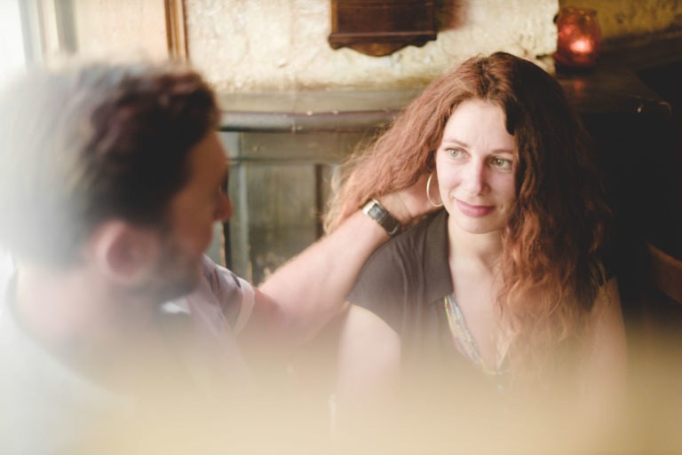 Une femme aux longs cheveux roux ondulés et à l'expression douce est assise à l'intérieur à côté d'un homme dont la main est doucement posée sur son épaule. L'arrière-plan est flou et aux tons chauds, comme le décor de Coup de foudre au café Brun Bordeaux, avec un panneau de bois et une petite bougie allumée sur une étagère, créant une atmosphère intime et chaleureuse.