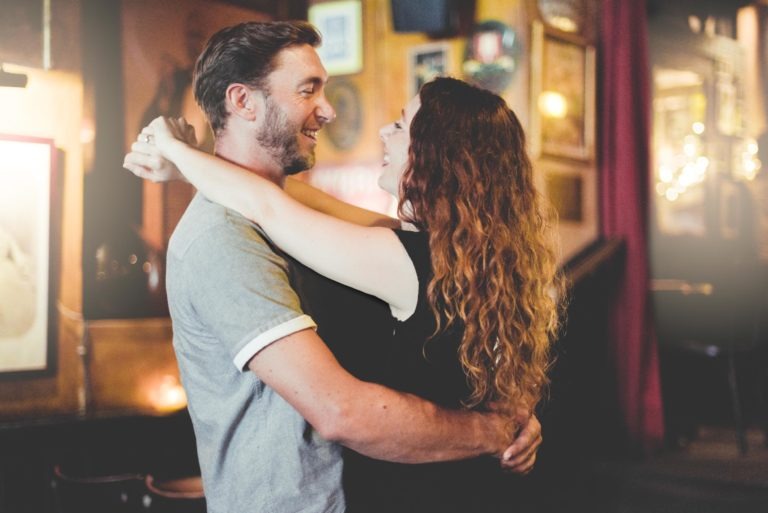 Un couple, sous le coup de foudre au café Brun Bordeaux, s'embrasse et se regarde chaleureusement dans une pièce confortable, à la lumière tamisée, décorée de tableaux encadrés et de lumières chaleureuses. L'homme porte un t-shirt gris et la femme a de longs cheveux bouclés dans un haut noir sans manches. Ils semblent heureux et profondément connectés.