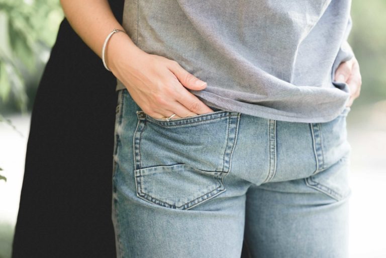 Gros plan d'une personne portant un jean bleu clair, les mains dans les poches et une chemise grise, rappelant un après-midi de détente au Café Brun Bordeaux. Le corps de la personne est légèrement tourné, ce qui montre les détails des poches et la texture du tissu. L'environnement comprend de la verdure floue en arrière-plan.