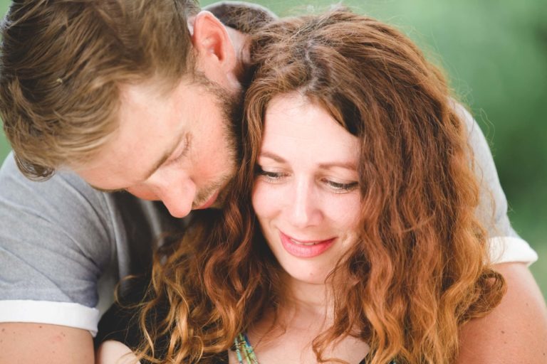 Un couple partage un moment de tendresse, l'homme posant doucement sa tête sur l'épaule de la femme. Tous deux ont les cheveux clairs ; l'homme a les cheveux courts tandis que la femme a les cheveux longs et bouclés. Ils sourient doucement, rappelant un coup de foudre au café Brun Bordeaux. L'arrière-plan est une verdure floue.