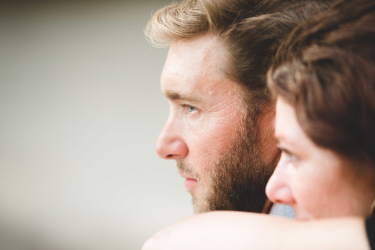 Gros plan de profil d'un homme et d'une femme aux tons de peau clairs. L'homme a les cheveux châtain clair et une barbe, le regard fixé au loin. La femme, partiellement cachée, a des cheveux bruns ondulés reposant sur son épaule. Leur pose dégage un air de « coup de foudre au café Brun Bordeaux », sur fond de flou doux et neutre.