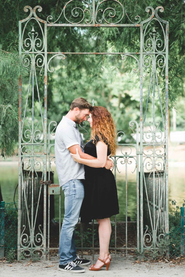 Un couple se tient debout sous une arche décorative en fer forgé, enlacés et en se touchant le front. L'homme, vêtu d'une chemise gris clair et d'un jean, fait face à la femme, qui porte une robe noire et des talons marron. Un feuillage vert les entoure, et un arrière-plan serein évoque un décor de jardin, une scène qui rappelle le coup de foudre au café Brun Bordeaux.