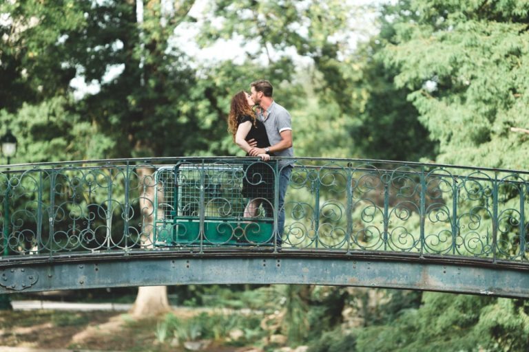 Un couple s'embrasse sur un pont métallique décoré entouré de verdure luxuriante. La femme a les cheveux bouclés et porte une robe noire, tandis que l'homme a les cheveux courts et porte une chemise gris clair. Leur étreinte fait écho au sentiment romantique de "Coup de foudre au café Brun Bordeaux", avec le paysage serein du parc en arrière-plan.