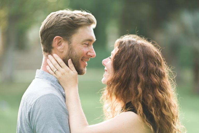 Un couple, un homme et une femme, se tiennent côte à côte à l'extérieur, souriant chaleureusement. La femme, aux cheveux longs et bouclés, tient doucement le visage de l'homme, mettant en valeur une bague de fiançailles à son doigt, un moment qui rappelle leur coup de foudre au café Brun Bordeaux. L'arrière-plan est un feuillage vert flou.