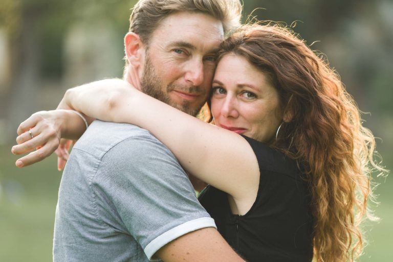 Un couple s'embrasse en plein air, la femme entourant les épaules de l'homme de ses bras. Ils regardent la caméra avec contentement, se rappelant peut-être leur coup de foudre au café Brun Bordeaux. L'homme a les cheveux châtains courts et clairs, une barbe et porte une chemise grise. La femme a les cheveux roux bouclés et longs et est vêtue d'un haut noir. L'arrière-plan est vert et flou.