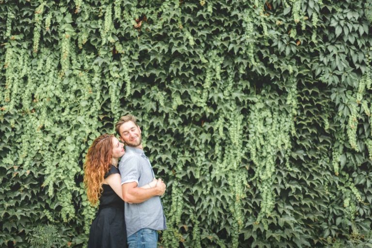 Un couple se tient devant un mur vert luxuriant recouvert de lierre. La femme rousse, vêtue d'une robe noire, serre l'homme blond dans ses bras par derrière, et tous deux sourient. Les feuilles texturées créent une toile de fond naturelle et vibrante pour cette pose affectueuse, rappelant leur Coup de foudre au café Brun Bordeaux.