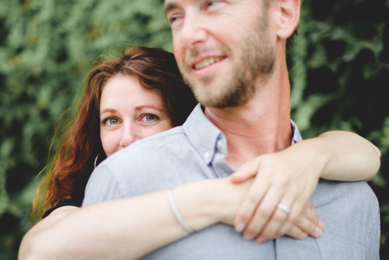 Une femme aux cheveux bruns enlace un homme par derrière, posant son menton sur son épaule. Elle a un sourire doux et regarde directement la caméra. L’homme, vêtu d’une chemise à col bleu clair, semble sourire et regarder légèrement vers la droite. C’est comme s’ils venaient de vivre un coup de foudre au café Brun Bordeaux. L’arrière-plan est flouté par un feuillage vert.