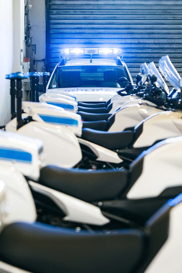 Un véhicule de la police municipale de Bordeaux avec des feux bleus est garé dans un garage. Devant lui, plusieurs motos de police blanches sont alignées. Le garage a une porte en tôle ondulée partiellement fermée en arrière-plan. La scène est bien éclairée, ce qui met en valeur le design épuré des véhicules.