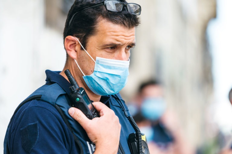 Un policier en uniforme de la police municipale de Bordeaux, portant un masque bleu et des lunettes sur la tête, tient un talkie-walkie près de sa bouche. L'arrière-plan est légèrement flou, montrant un autre individu masqué dans un uniforme similaire, indiquant une situation de sécurité publique ou de maintien de l'ordre.