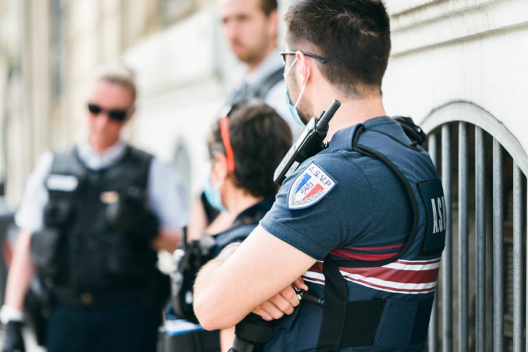 Un groupe de quatre policiers, membres de la police municipale de Bordeaux, se tient près d'un bâtiment blanc. Trois d'entre eux portent des gilets noirs, tandis qu'un autre porte un gilet bleu marine à rayures rouges et blanches et l'inscription "ASVP" sur l'épaule. Ils semblent être en conversation tandis qu'un des policiers s'appuie contre le bâtiment.