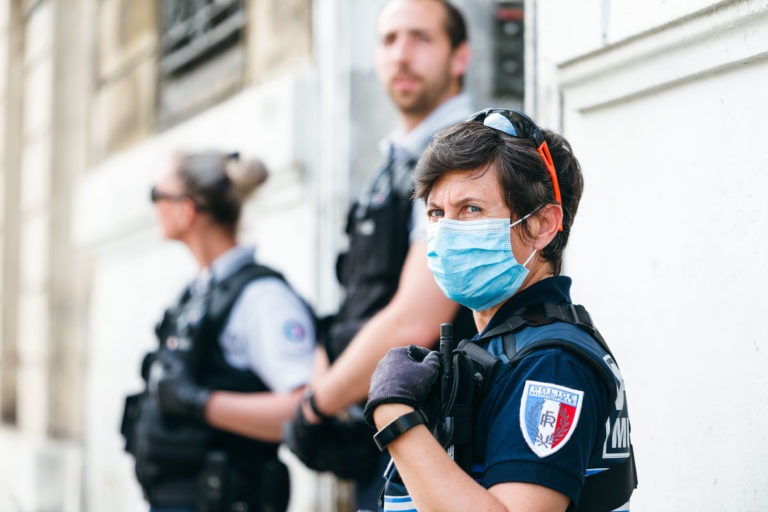 Un membre de la police municipale de Bordeaux en uniforme bleu et cagoule, les cheveux courts et des lunettes de soleil sur la tête, se tient au premier plan, regardant vers la droite. Ils sont entourés de deux collègues flous en uniformes similaires à l'arrière-plan. Le décor semble être à l'extérieur avec des murs de couleur claire.