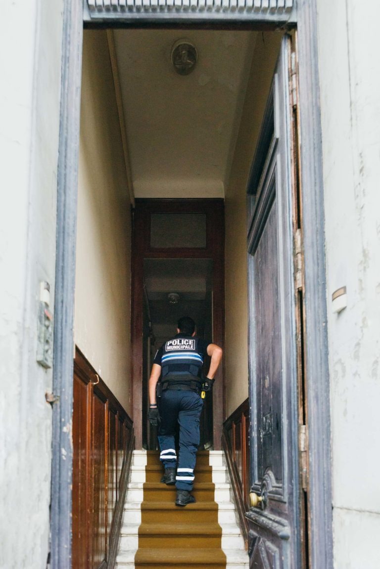 Un policier de la Police Municipale de Bordeaux monte un escalier étroit et faiblement éclairé à l'intérieur d'un vieux bâtiment. Il porte un uniforme sombre avec le mot « Police » écrit au dos. L'escalier est encadré par des panneaux en bois et une porte entrouverte, ce qui laisse penser qu'il s'agit d'un bâtiment résidentiel ou d'un petit local commercial.