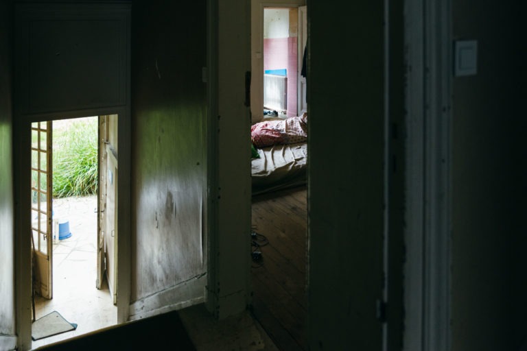 Un couloir faiblement éclairé mène à une pièce partiellement ouverte avec un lit en désordre recouvert de couvertures et d'oreillers. Une lumière douce filtre par une fenêtre, illuminant les murs usés et les objets éparpillés sur le sol. La présence de la police municipale de Bordeaux suggère une enquête, car une porte au bout du couloir s'ouvre sur un jardin envahi par la végétation.