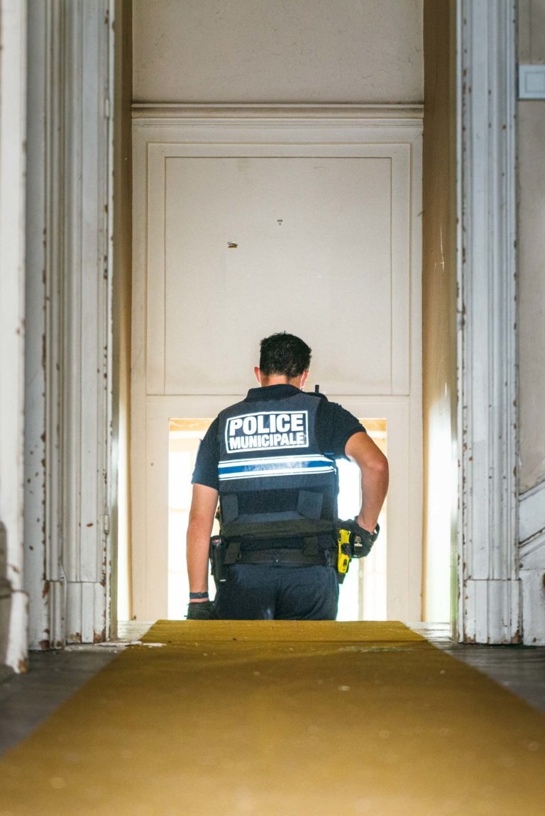 Un policier en uniforme de la « Police Municipale de Bordeaux » se tient dans un couloir faiblement éclairé, le dos tourné. La ceinture de l'agent contient divers outils et équipements, dont un pistolet paralysant. Le couloir est doté de murs lambrissés de blanc et d'un tapis jaune menant à une porte ouverte traversée par la lumière du soleil.