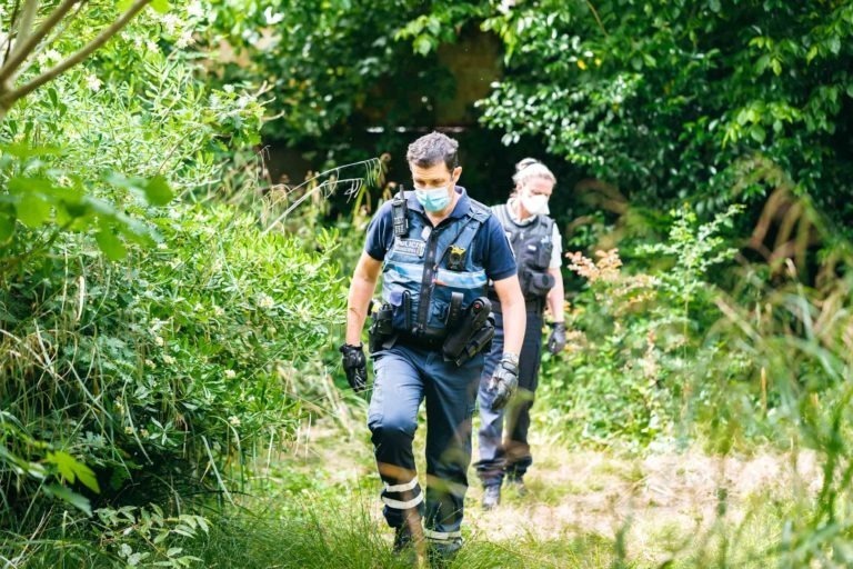 Deux policiers de la Police Municipale de Bordeaux, portant des masques, marchent dans un espace vert luxuriant au feuillage dense. Ils sont vêtus d'uniformes sombres et suivent un étroit sentier de terre, entouré d'herbes hautes et d'arbustes. La scène semble être une patrouille de jour dans une zone boisée ou un parc.