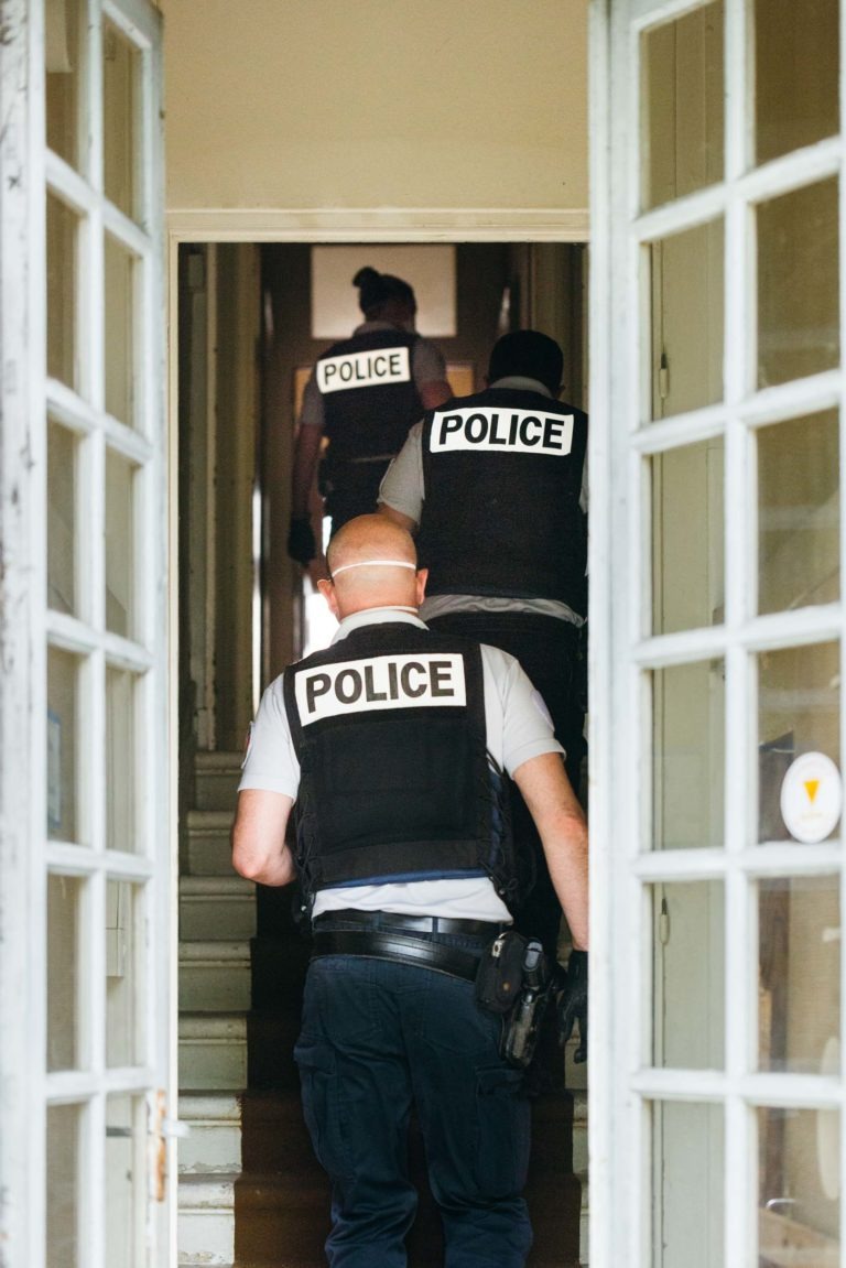 Trois policiers municipaux de Bordeaux en uniforme, portant chacun un gilet avec l'inscription « POLICE », montent un escalier à l'intérieur d'un bâtiment. Le cadre de la porte par laquelle ils passent est ouvert, révélant le couloir intérieur et les marches menant au sommet. La scène est bien éclairée, ce qui indique qu'il fait peut-être jour.