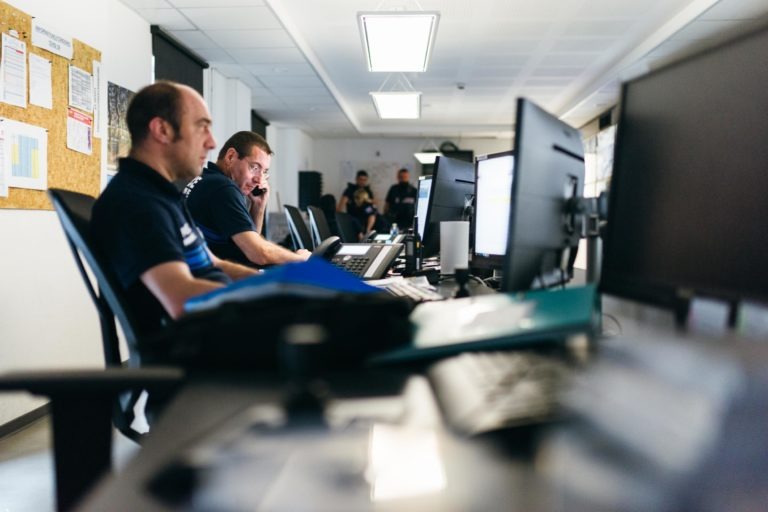 Dans une pièce très éclairée, probablement celle de la police municipale de Bordeaux, deux hommes sont assis à leur bureau, concentrés sur leurs écrans d'ordinateur. L'un d'eux utilise un téléphone. La pièce est équipée d'un tableau d'affichage sur lequel sont épinglés des documents à gauche et d'un tableau blanc ou d'un écran au fond. D'autres personnes et du matériel de bureau sont visibles en arrière-plan.