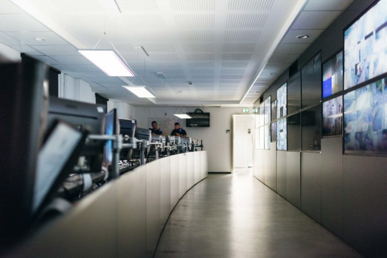 Une salle de contrôle moderne est équipée de plusieurs écrans et postes de travail avec des ordinateurs disposés le long d'un bureau incurvé. Deux personnes de la police municipale de Bordeaux sont visibles en arrière-plan, en pleine conversation. Plusieurs grands écrans affichant diverses données et images bordent le mur de droite. La salle est bien éclairée par des plafonniers.