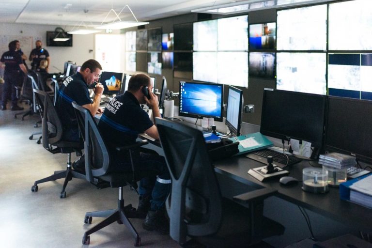 Une salle de contrôle moderne avec plusieurs écrans diffuse des images de surveillance. Deux hommes en uniforme noir sont assis à leur bureau, concentrés sur leurs écrans. Autour d'eux se trouvent d'autres postes informatiques et du matériel de bureau. L'arrière-plan montre des agents de la police municipale de Bordeaux et des écrans supplémentaires fixés au mur.