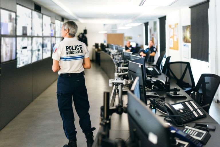 Une personne en uniforme de la « Police Municipale de Bordeaux » se tient dans une salle de contrôle, surveillant plusieurs écrans diffusant des images de surveillance. La salle est équipée de plusieurs bureaux équipés d'ordinateurs et d'appareils de communication. Deux autres agents sont assis à des bureaux en arrière-plan, également occupés à la surveillance.