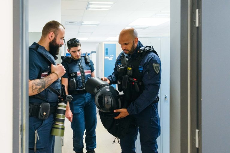 Trois policiers en uniforme de la police municipale de Bordeaux sont réunis dans ce qui semble être un vestiaire. L'agent de droite tient un grand équipement ressemblant à un casque et le montre aux deux autres. Les agents ont l'air sérieux et discutent.