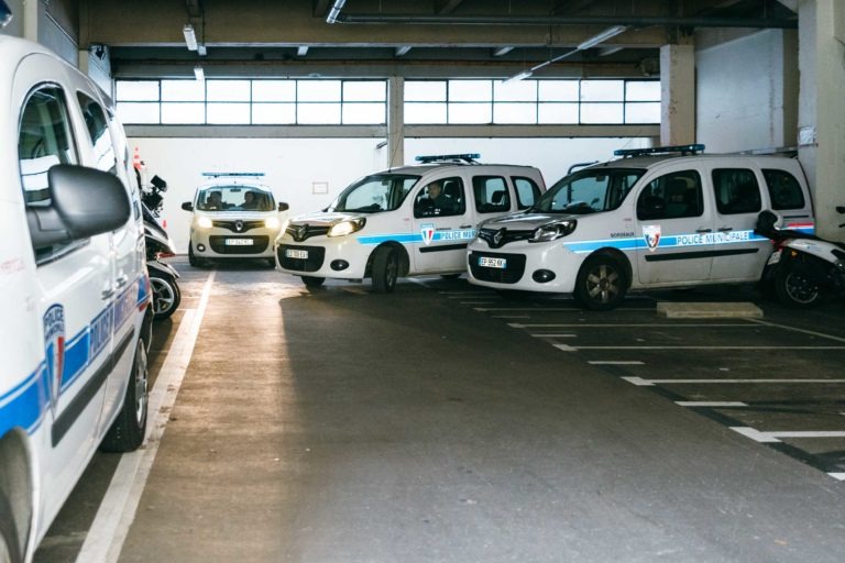 Un parking couvert, faiblement éclairé, abrite plusieurs véhicules de police blancs disposés à divers endroits. Chaque véhicule, marqué de bandes bleues et de l'emblème de la Police Municipale de Bordeaux, est prêt à l'action. Le garage est doté de murs en béton, de hautes fenêtres laissant entrer la lumière naturelle et de voies de stationnement marquées au sol.