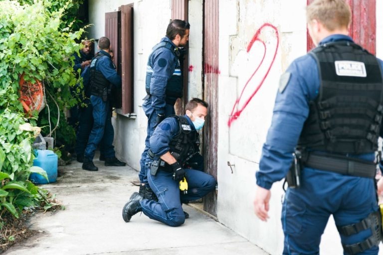 Un groupe de policiers municipaux de Bordeaux, vêtus d'uniformes bleu marine et d'équipements tactiques, mènent une opération de recherche. Un policier est à genoux et semble examiner ou travailler sur une porte. Le bâtiment est ancien et un grand numéro "2" rouge est peint à la bombe sur le mur. Un feuillage vert entoure la zone.