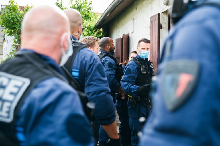 Un groupe de policiers municipaux de Bordeaux, vêtus d'une tenue tactique, sont rassemblés devant un bâtiment aux volets marron. Tous portent des masques et certains ont le crâne visiblement rasé. Un feuillage vert est visible en arrière-plan. L'atmosphère semble tendue et concentrée.