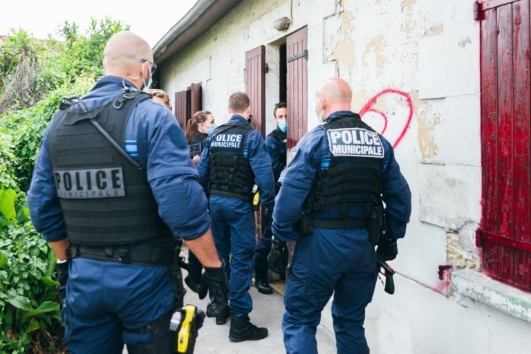 Un groupe de cinq policiers municipaux, vus de dos, portant des uniformes bleus et des gilets noirs marqués « POLICE MUNICIPALE DE BORDEAUX », sont rassemblés devant un bâtiment blanc patiné aux volets marron. Un policier entre par une porte. Des graffitis, dont un cercle rouge, sont visibles sur le mur.