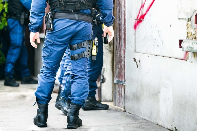 Des policiers de la police municipale de Bordeaux, vêtus d'uniformes bleus et d'équipements tactiques, se tiennent dans une ruelle à côté d'un bâtiment délabré parsemé de graffitis. L'arme et l'équipement d'un policier sont visibles. La scène semble tendue, suggérant qu'une opération policière ou une enquête est en cours.