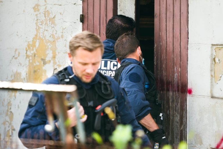 Trois agents de la police municipale de Bordeaux, vêtus d'uniformes bleus et d'équipements tactiques, pénètrent dans un bâtiment délabré dont la peinture s'écaille. L'un des agents au premier plan tient un outil tandis que les deux autres, partiellement visibles, se concentrent sur la porte d'entrée. La scène suggère une enquête ou une intervention active.