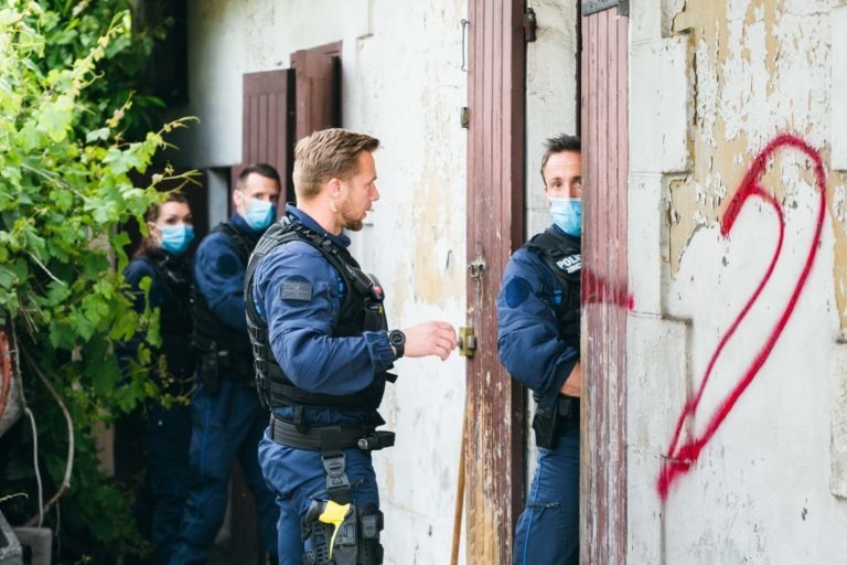 Quatre agents de la police municipale de Bordeaux, portant des uniformes bleus et des masques, effectuent une perquisition dans un vieux bâtiment délabré. Un agent ouvre une porte tandis qu'un autre regarde à l'intérieur. Le bâtiment est partiellement recouvert de feuillage et un grand graffiti rouge « 2 » est peint sur le mur extérieur.