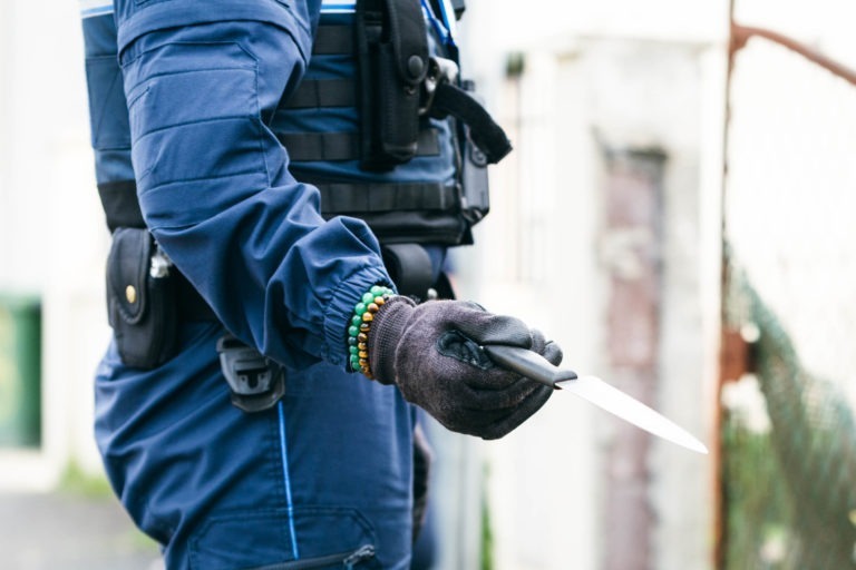 Un membre de la police municipale de Bordeaux, vêtu d'un uniforme bleu et de gants noirs, tient un couteau dont la lame est pointée vers le bas. L'uniforme comporte de nombreux compartiments et accessoires. L'arrière-plan suggère un décor extérieur avec des bâtiments et des clôtures flous, tandis que le visage de la personne reste invisible.