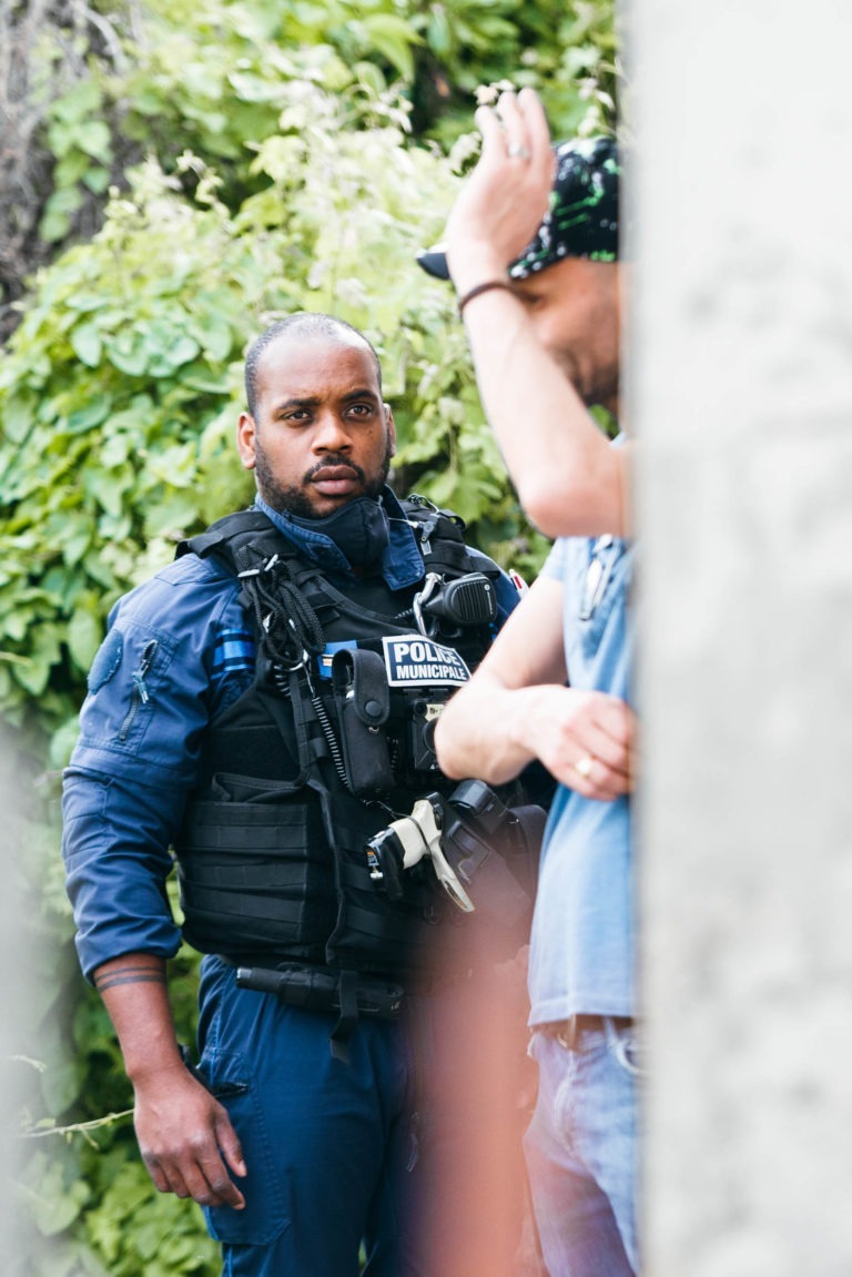 Un policier en tenue tactique avec un gilet « POLIZEI MUNICH POLICE » regarde vers la caméra. Il se tient près d'une personne en chemise bleue et chapeau de camouflage, qui lève le bras. L'arrière-plan, luxuriant de feuillage vert, contraste avec l'intensité souvent observée dans les interactions de la police municipale de Bordeaux.