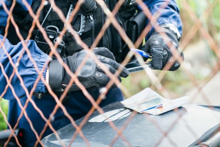 Une personne portant des gants tactiques, un gilet pare-balles et un uniforme bleu de la police municipale de Bordeaux est aperçue à travers une clôture grillagée rouillée. Elle inspecte un petit appareil électronique fin avec une pince. Un bloc-notes et d'autres objets reposent sur une boîte noire devant elle, suggérant un contexte de maintien de l'ordre.