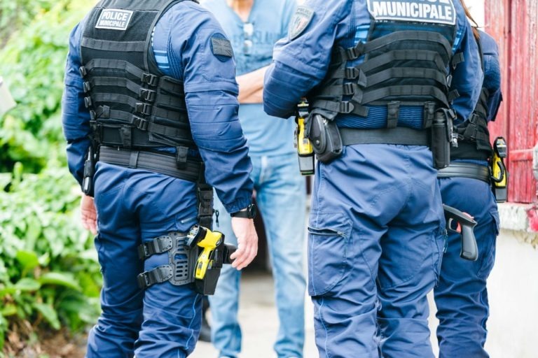 Trois policiers en uniforme bleu avec l'inscription « POLICE MUNICIPALE DE BORDEAUX » sur leur gilet se tiennent debout face à une personne en tenue décontractée. Les policiers portent des équipements tels que des menottes et des tasers attachés à leur gilet. De la verdure est visible en arrière-plan, suggérant un décor extérieur.