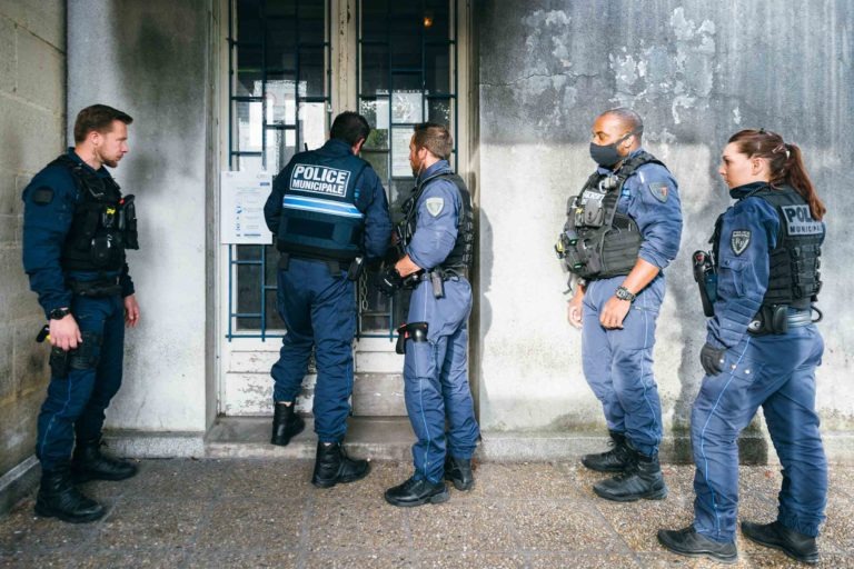 Cinq agents de la police municipale de Bordeaux se tiennent devant l'entrée d'un bâtiment, tous en uniforme. L'un d'eux se concentre sur le déverrouillage d'une porte métallique tandis que les autres observent et se tiennent prêts à aider. Les agents portent un équipement tactique, notamment des gilets étiquetés « Police Municipale ».