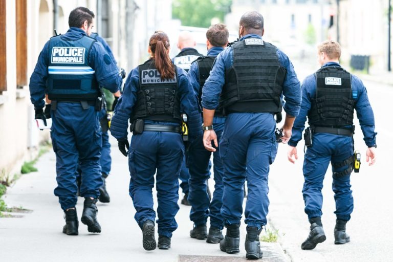 Un groupe de six policiers municipaux, cinq hommes et une femme, s'éloignent sur un trottoir d'une ville. Ils portent des uniformes bleu foncé et des gilets tactiques noirs étiquetés « POLICE MUNICIPALE DE BORDEAUX » en lettres blanches. L'arrière-plan montre des bâtiments et de la verdure.