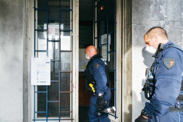 Deux agents en uniforme de la police municipale de Bordeaux, portant des masques et des équipements de sécurité, dont des gilets pare-balles et des ceintures utilitaires, pénètrent dans un bâtiment par une porte vitrée à cadre métallique. L'un des agents porte une lampe de poche. La porte est ornée d'affiches et l'extérieur du bâtiment semble vieilli et usé.