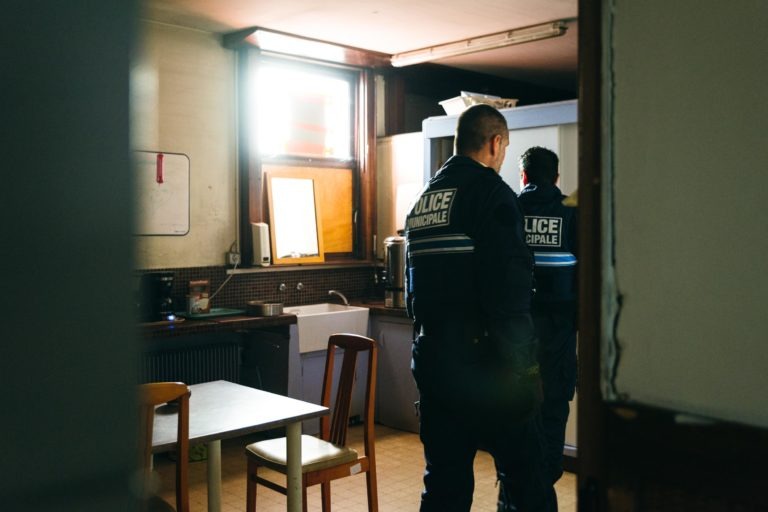 Deux policiers en uniforme sombre avec l'inscription « Police Municipale de Bordeaux » dans le dos sont vus dans une petite pièce faiblement éclairée avec un mobilier minimaliste comprenant une table, des chaises et du matériel de cuisine. Les policiers, détournés de la caméra, semblent concentrés sur quelque chose en dehors du cadre. La lumière naturelle provient d'une fenêtre.