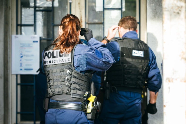 Deux policiers municipaux de Bordeaux en uniforme sont vus de dos, devant un bâtiment. Ils portent tous deux des gilets pare-balles et ont divers équipements attachés à leur ceinture. L'agent de gauche ajuste ses cheveux, tandis que l'agent de droite ajuste son masque facial.