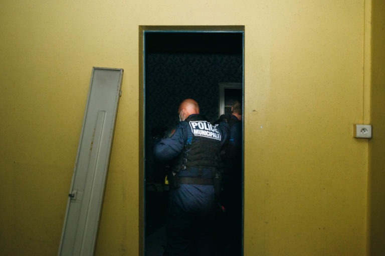 Deux policiers en tenue tactique, identifiés par « POLICE » au dos de leur gilet, pénètrent dans une pièce faiblement éclairée par une porte jaune. L'agent au premier plan appartient à la police municipale de Bordeaux. Une porte démontée est adossée au mur à gauche de la porte.