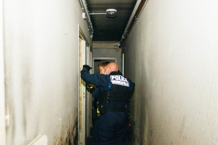 Deux policiers municipaux de Bordeaux, en uniforme, se tiennent dans un couloir étroit et mal éclairé aux murs blancs. L'un d'eux tient une lampe de poche, éclairant le chemin, tandis qu'ils regardent à travers une porte ouverte. Les murs montrent des signes d'usure et des tuyaux courent le long du plafond.