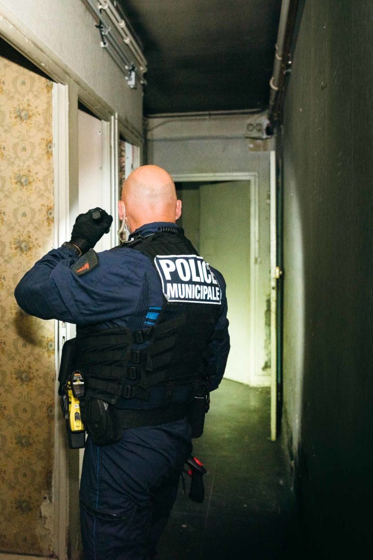 Un policier portant l'inscription « POLICE MUNICIPALE » dans le dos se tient dans un couloir étroit et mal éclairé à l'intérieur d'un bâtiment. L'agent, qui fait partie de la police municipale de Bordeaux, porte un uniforme bleu foncé et un gilet tactique et utilise une lampe de poche pour inspecter une pièce. Le couloir est recouvert de papier peint usé et la peinture s'écaille.