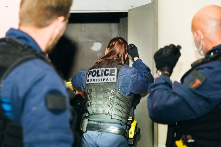 Plusieurs policiers en uniforme bleu et gilet tactique, dont l'un porte l'inscription "Police Municipale de Bordeaux" au dos, se trouvent dans un bâtiment aux murs décolorés. Ils semblent mener une perquisition ou une opération, l'un d'eux examinant de près ou pénétrant dans une porte tandis que d'autres se tiennent à proximité.