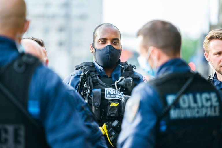 Cinq agents de la police municipale de Bordeaux se tiennent debout à l'extérieur, en pleine conversation. Vêtus d'uniformes bleus, de gilets pare-balles et de masques, ils dégagent un sens du devoir. Le personnage central regarde directement la caméra tandis que les autres discutent. Un décor urbain flou leur sert de toile de fond.